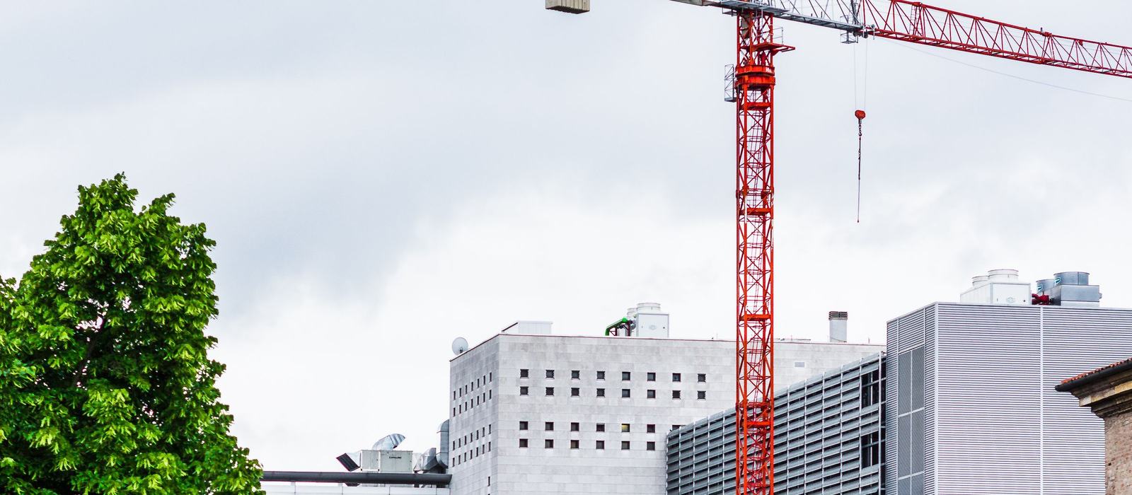 A red tower crane rising between office buildings and trees on an urban building site