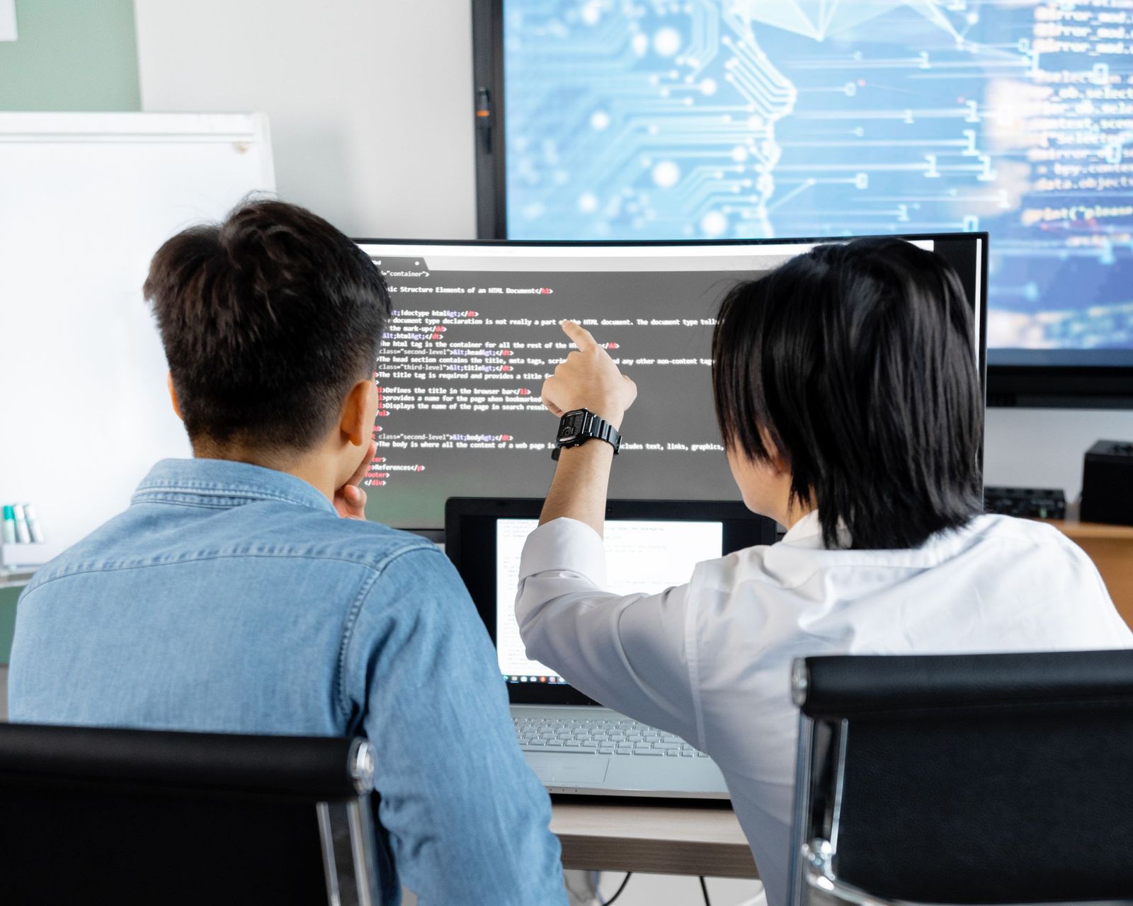 Two colleagues seen from behind at a workstation, one pointing at a line on the screen while the other follows
